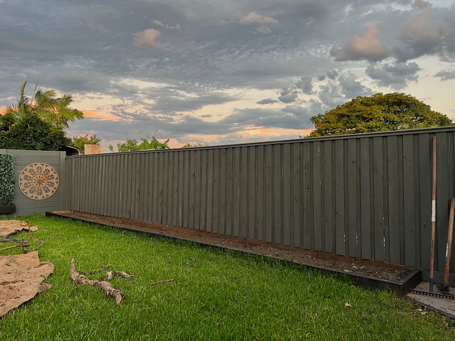 grass area in front of 10 meter long empty garden plot filled with soil in front of a fence, cloudy sky above