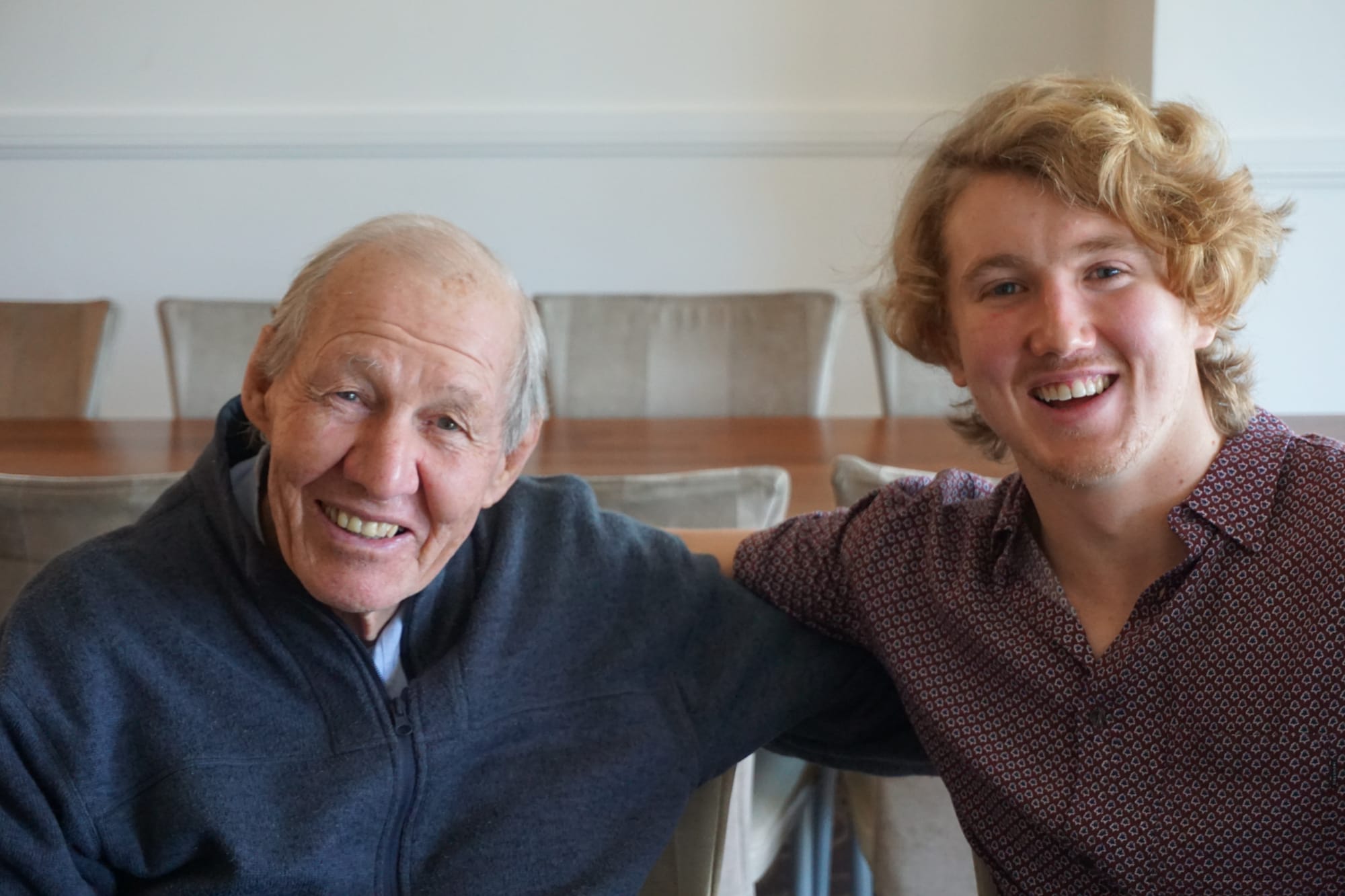 Two men sitting at a table, older man on left smiling and younger man on right smiling. Both are holding each other around the back.