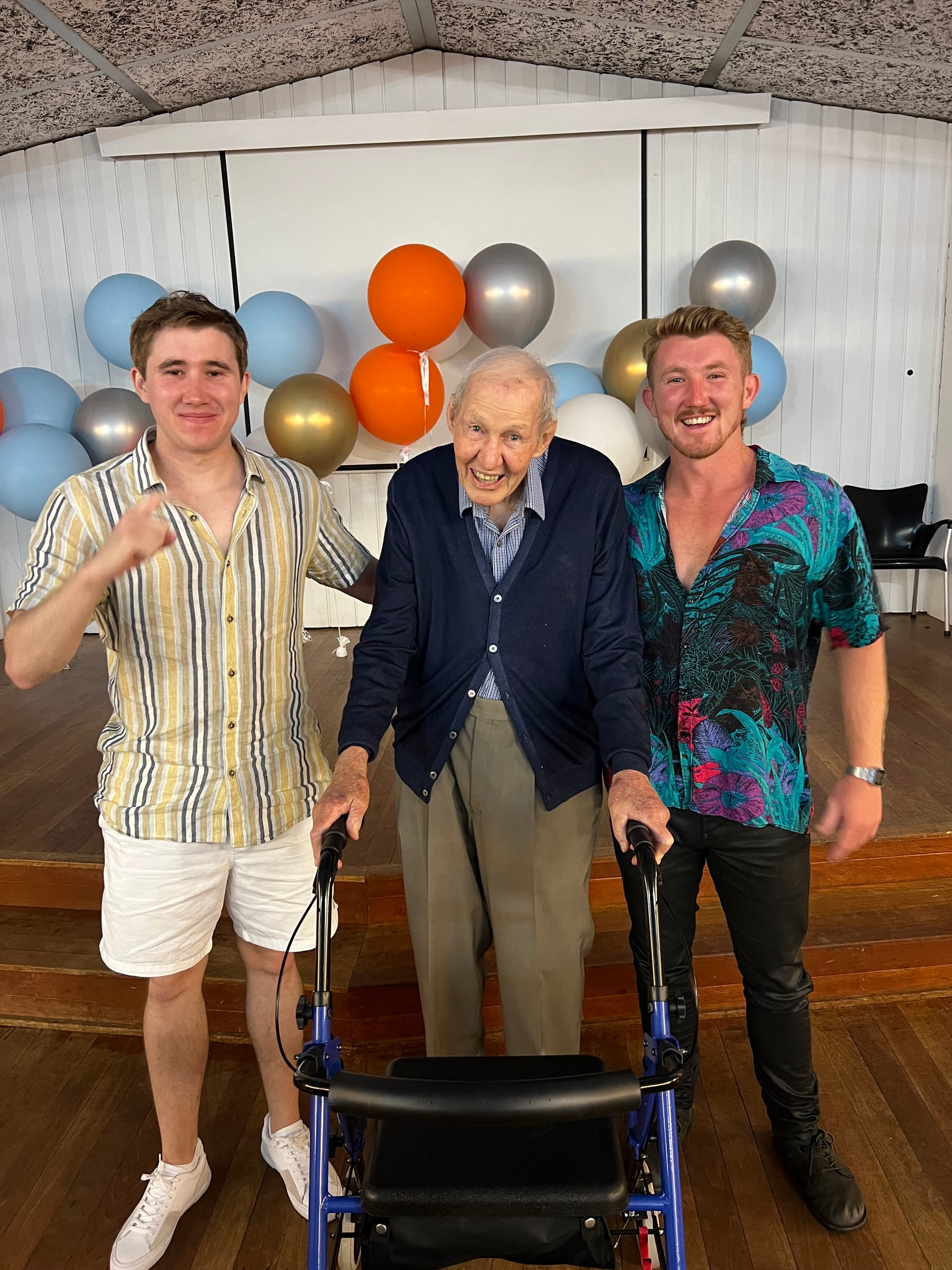 Three people standing in front of balloons. Two younger men on the outer sides and an older gentlemen in the middle with a walking frame.